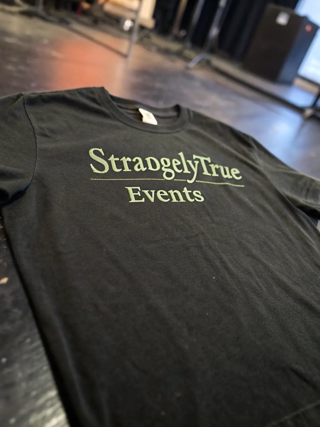 A close-up shot of a black t-shirt with the Strangely True Events logo in military green, subtly placed on the chest. The background is a blurred, dark room with a hint of mystery.